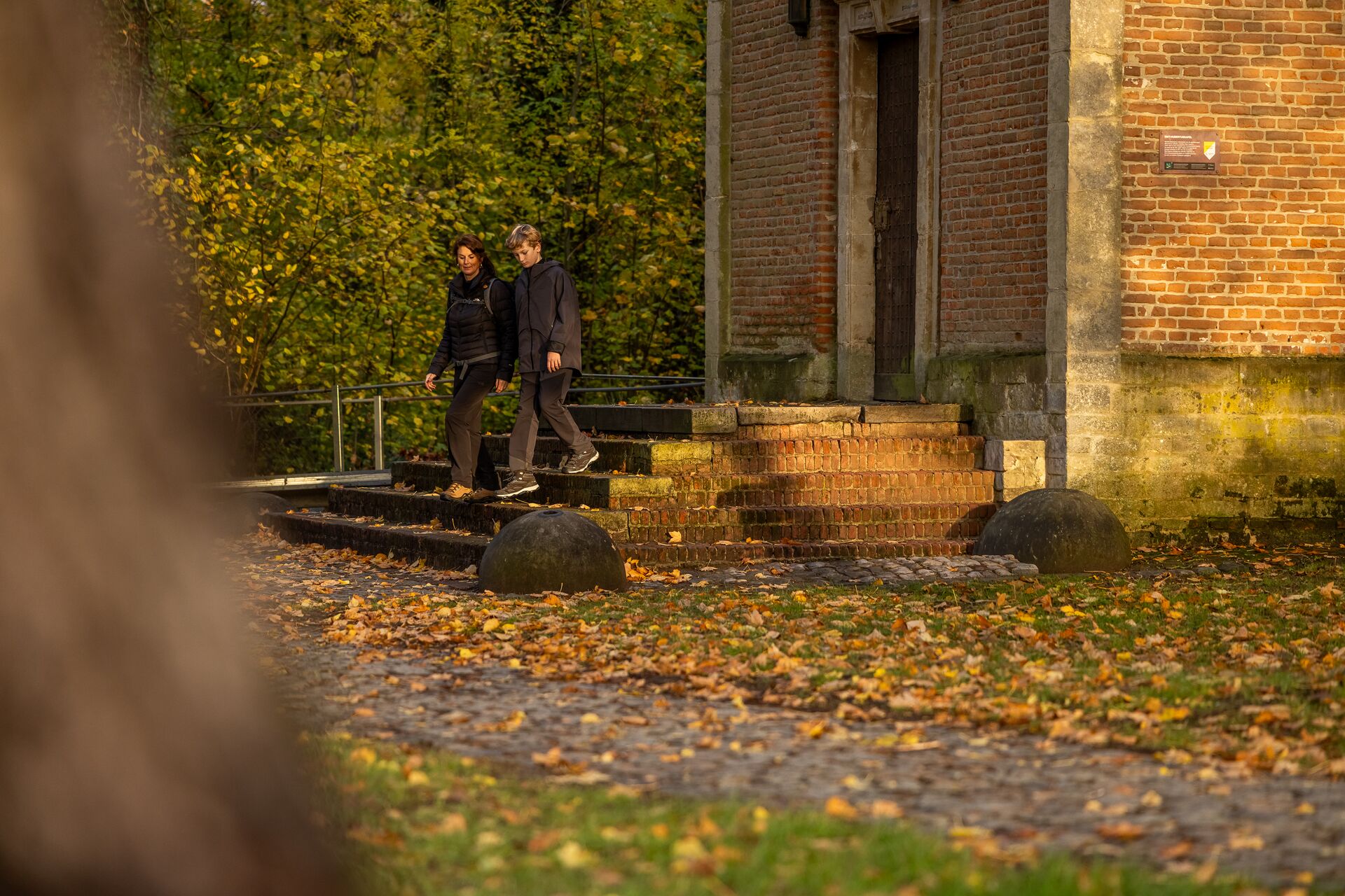 Due persone scendono i gradini di pietra di un edificio in mattoni circondato da fogliame autunnale. La calda luce del sole e le foglie cadute creano un’atmosfera tranquilla e serena.