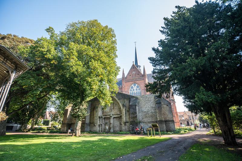 Historisch gebouw met een hoge torenspits, omgeven door weelderige groene bomen op een zonnige dag. Een kronkelend pad loopt door het goed onderhouden grasgebied.