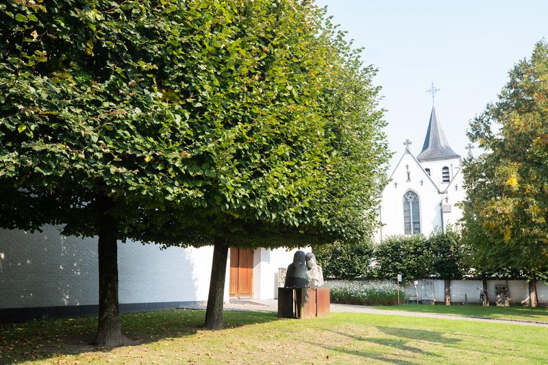 Un cortile tranquillo presenta un grande albero che fa ombra a una panchina con due sculture sedute. Una chiesa bianca con un alto campanile si trova sullo sfondo.