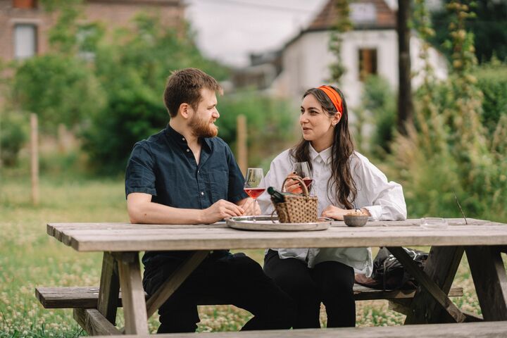 Een man en een vrouw zitten aan een picknicktafel in een tuin en genieten van drankjes. Ze voeren een vriendelijk gesprek, wat een ontspannen en aangename sfeer oproept.