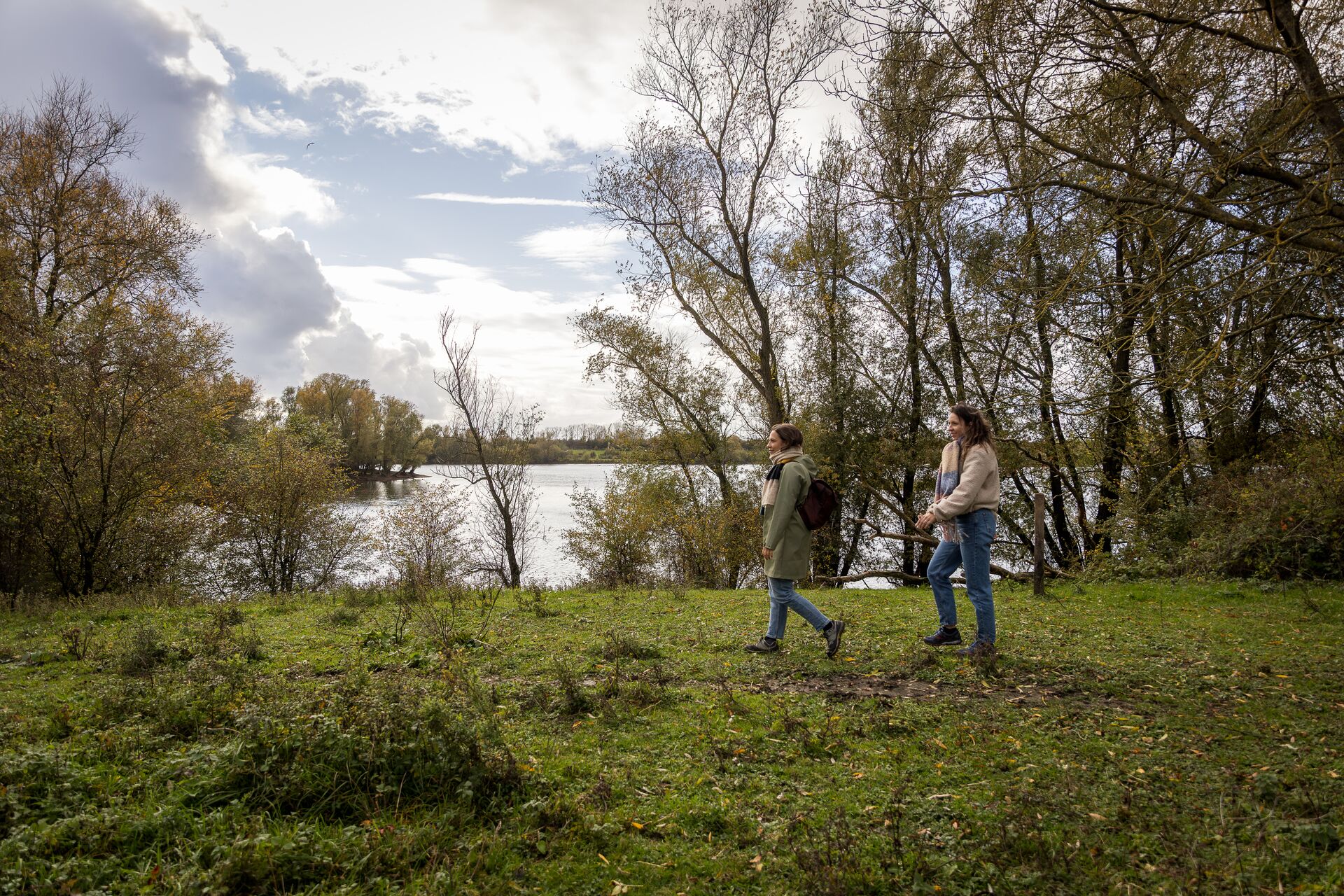 Dos personas caminan por un sendero de hierba junto a un lago tranquilo, rodeados de árboles otoñales bajo un cielo parcialmente nublado, evocando paz y relajación.