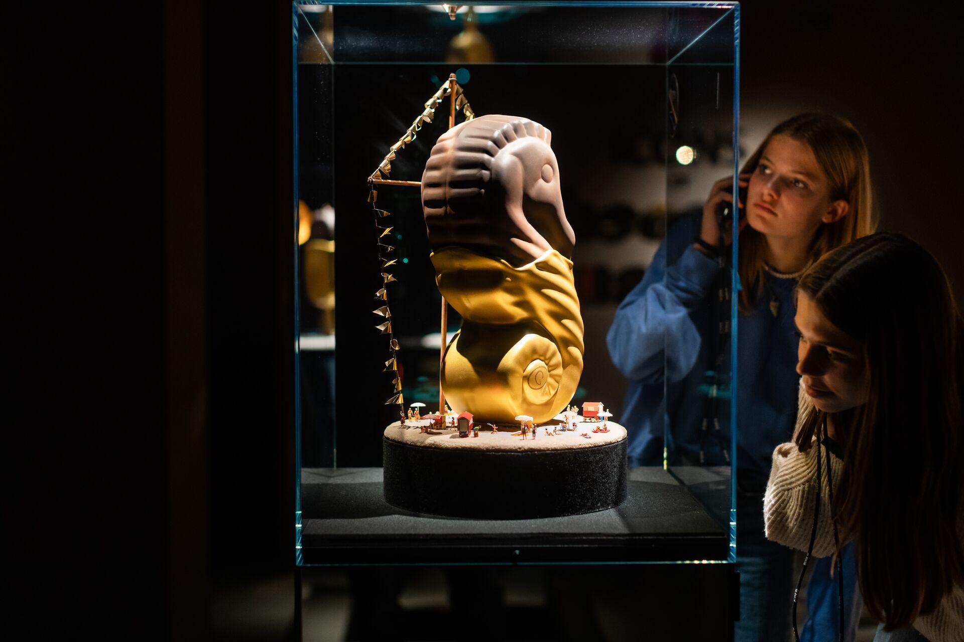 Chocolate Nation - Girls view sculpture in case Two girls look at a chocolate sculpture in a glass display case with a large snail-like head and miniature figures.