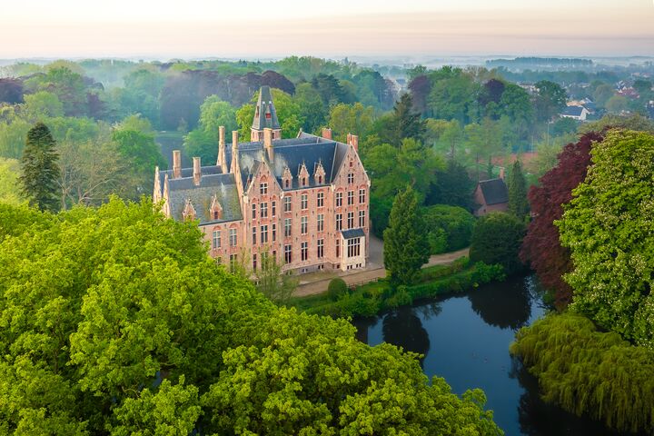 Schloss Loppem Luftaufnahme eines historischen Schlosses, umgeben von üppigen grünen Bäumen und Gärten. Ein ruhiger Teich spiegelt das lebendige Laub wider und schafft eine friedliche, malerische Szene.