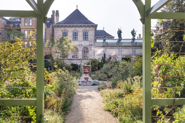 Rubenshuis Een weelderige tuin, omlijst door een boog, leidt naar een groot, historisch gebouw. De scène is sereen, met levendig groen en een helderblauwe lucht erboven.