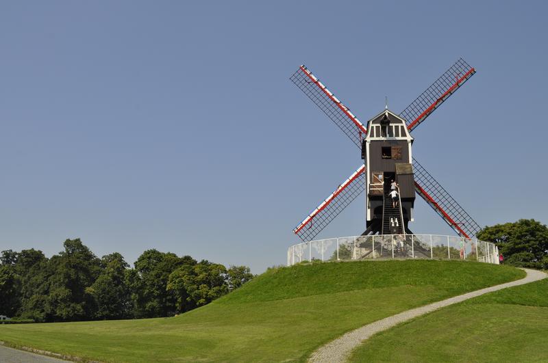 A traditional windmill with red and white blades stands atop a grassy hill under a clear blue sky. A winding path leads up to it, conveying serenity.