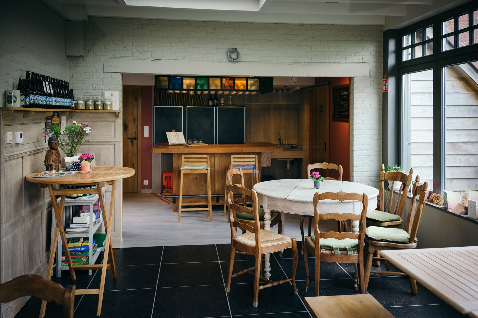 Erlebe die Hausbrauerei Karrelees Gemütliches Café-Interieur mit Holztischen und Stühlen. Ein runder Tisch ist mit kleinen Blumen geschmückt. Regale zeigen Flaschen. Eine warme, einladende Atmosphäre.