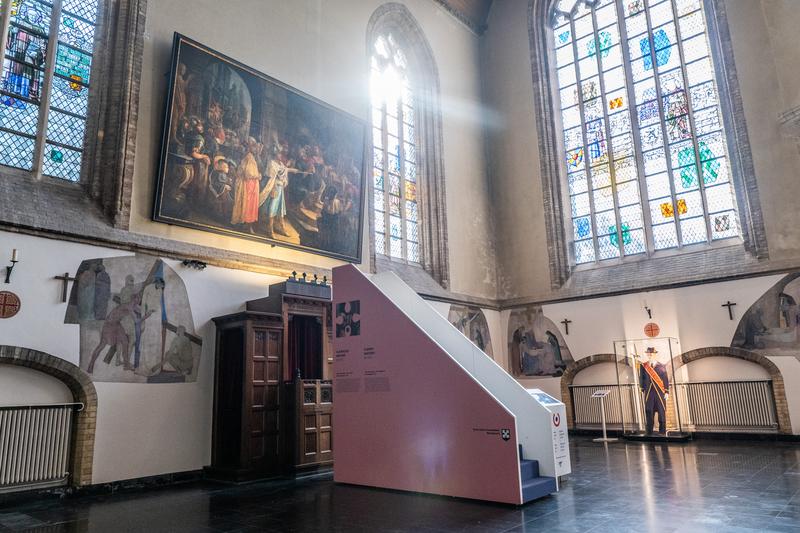 Een door de zon verlichte ruimte in een historisch gebouw met grote glas-in-loodramen, een levendig fresco en een koninklijke uniformtentoonstelling, wat een majestueuze sfeer creëert.