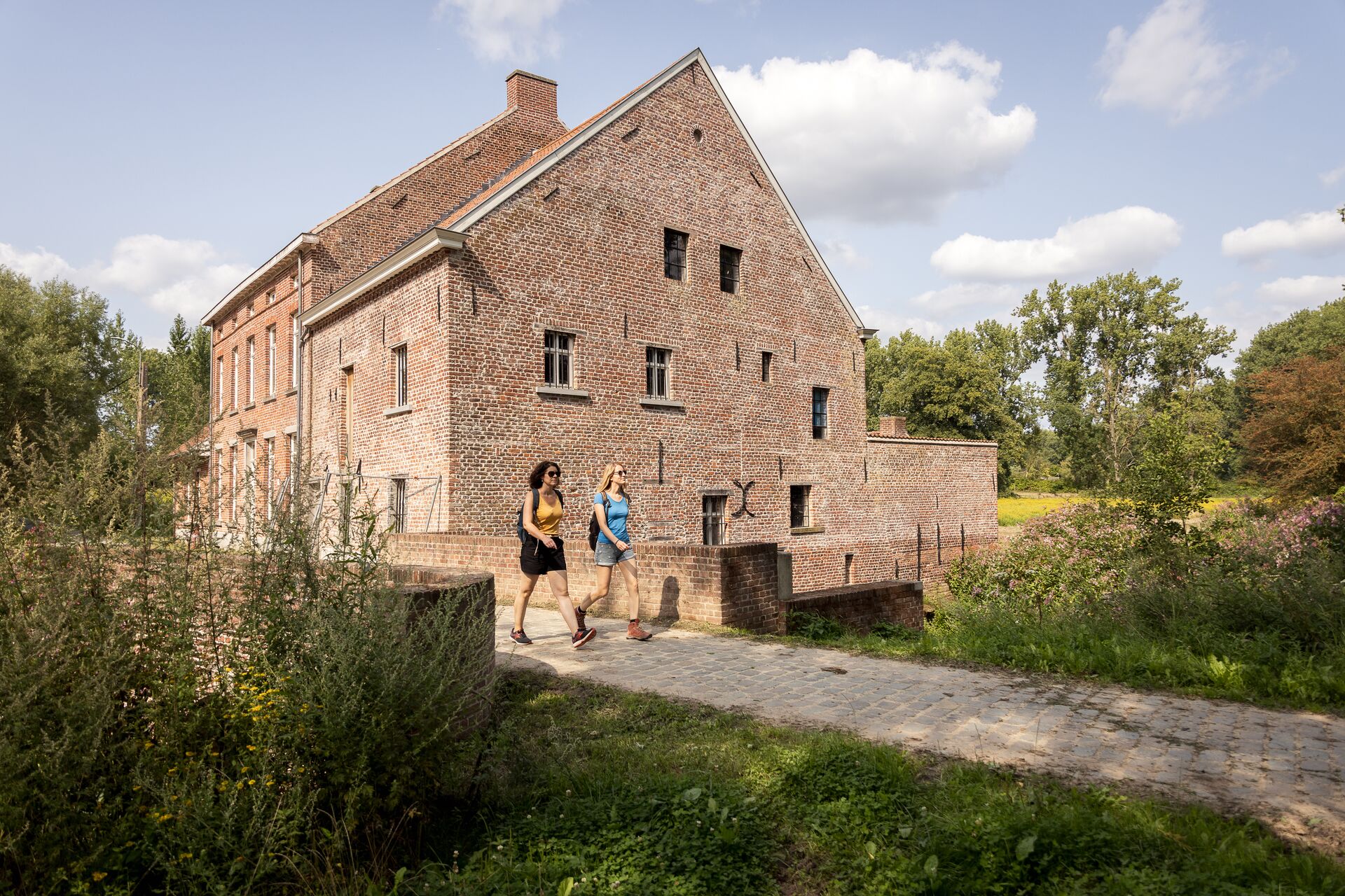 Due persone camminano su un sentiero acciottolato accanto a un edificio storico in mattoni, circondato da una vegetazione rigogliosa. La scena è tranquilla sotto un cielo parzialmente nuvoloso.