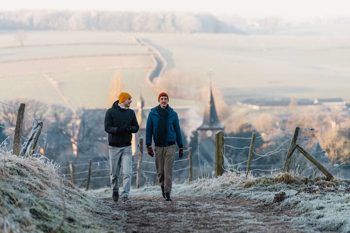 Two people wearing winter jackets walk along a frosty rural path. The landscape is cold and misty, with fields and trees covered in frost, evoking a serene, tranquil mood.