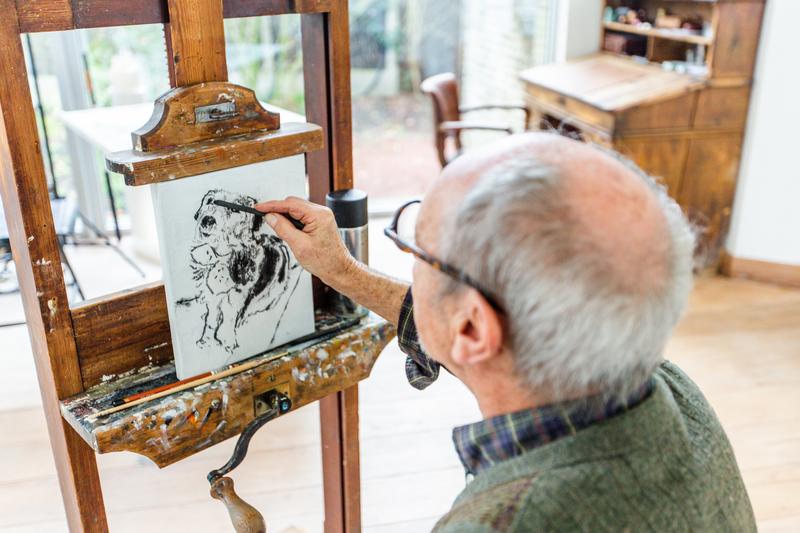 An elderly man in glasses sketches a dog on a canvas using charcoal. Sunlit room with wooden furniture in the background. Focused and peaceful ambiance.