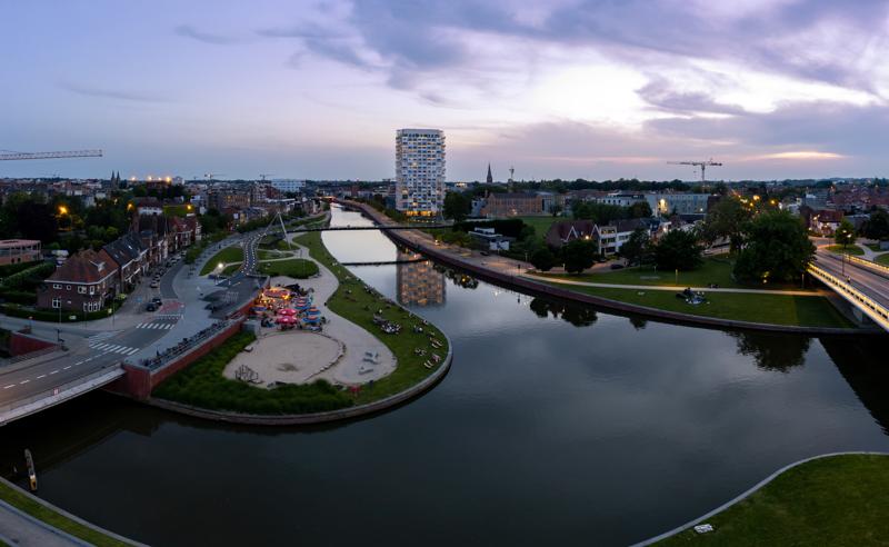 Luchtfoto van een sereen stadsbeeld aan de rivier bij schemering, met een reflecterende rivier, een zandstrand en een hoog, modern gebouw.