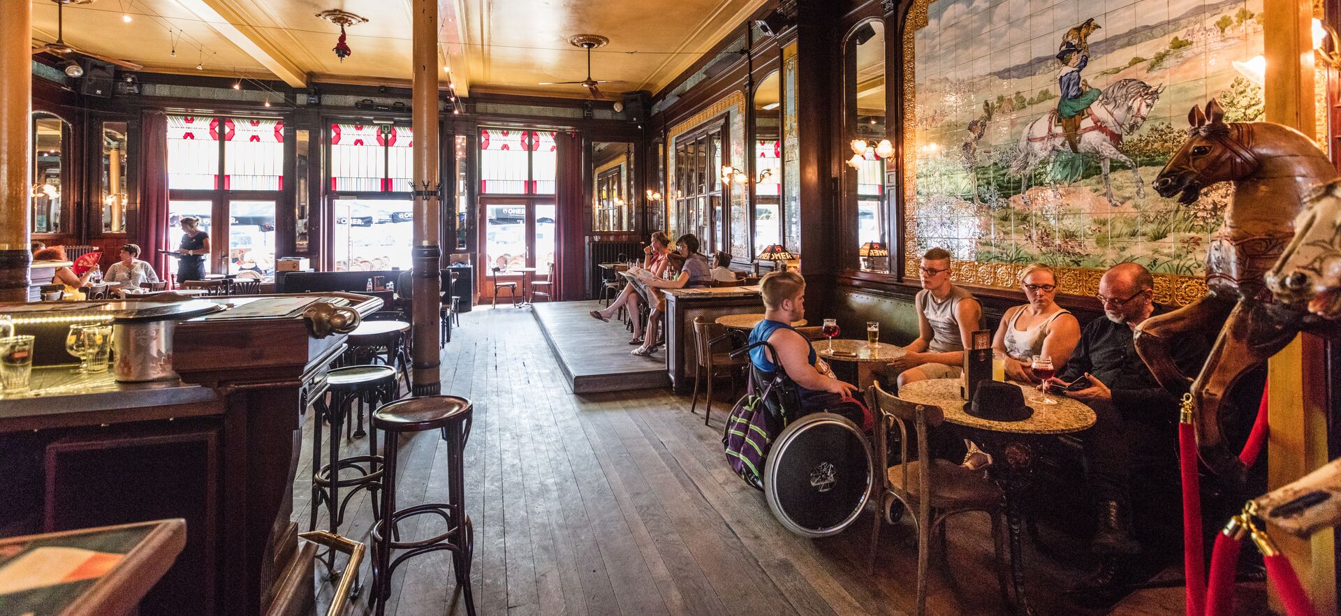 Café interior with guests and bar Interior of a café with bar, wooden floor and guests at tables; a man in a wheelchair sits in front.