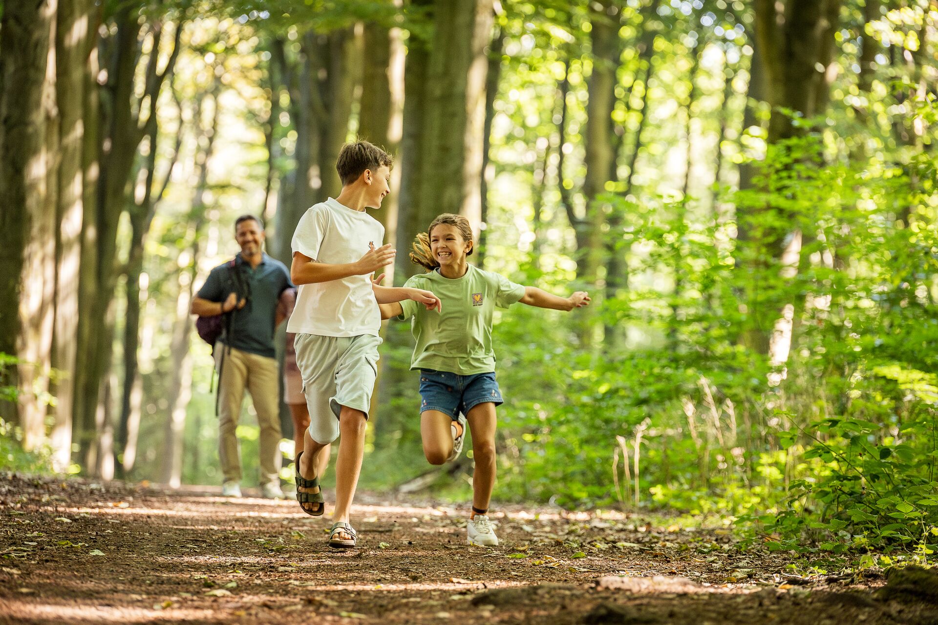 Een jongen en een meisje rennen vrolijk over een zonovergoten bospad, omgeven door groene bomen. Twee volwassenen lopen achter hen en glimlachen, wat een levendig gezinsmoment creëert.