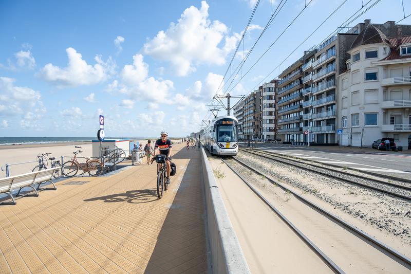 Cyclist on the seafront promenade with beach on the left and the coastal tram beside apartment buildings.
