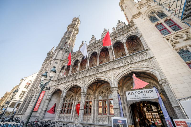 Gothic-style building with arches and ornate details, flying red flags, housing the Historium Bruges. Clear sky enhances the historic and grand atmosphere.