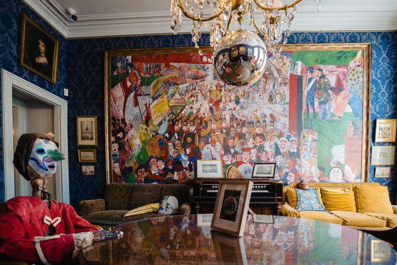 Een levendige kamer met een groot, kleurrijk muurschildering die een drukke scène toont. Een reflecterende kroonluchter hangt boven pluche gele banken, poppen en ingelijste foto’s. De setting is artistiek en eclectisch.