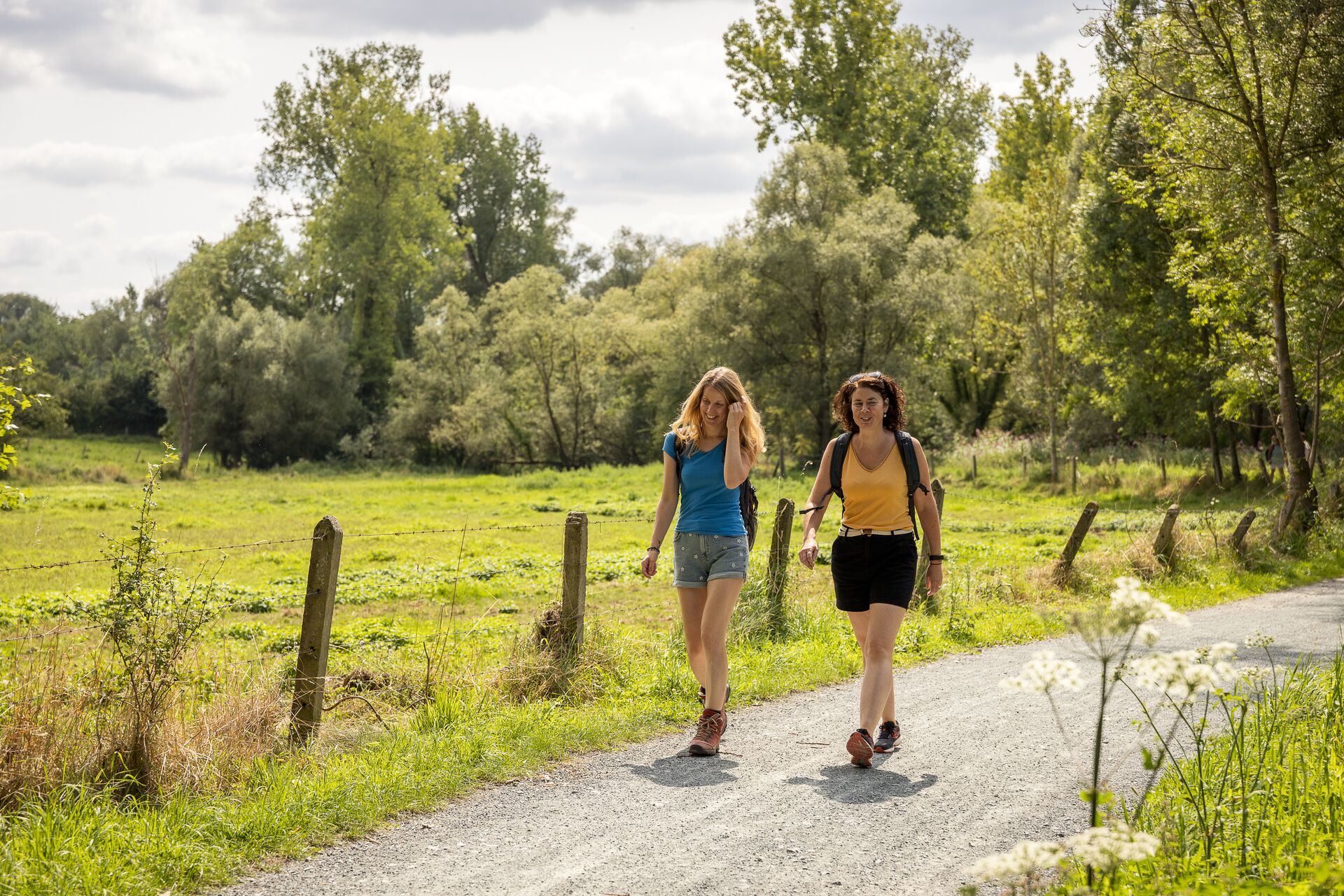 Due donne camminano su un sentiero ghiaioso soleggiato accanto a un campo verde e rigoglioso, circondate da alberi. Appaiono rilassate e si godono una piacevole giornata all’aperto.