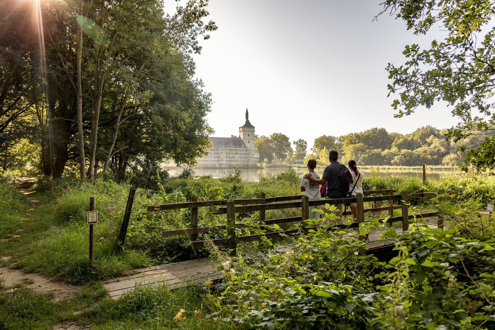 Drie personen staan op een houten brug in een weelderig park, kijkend naar een verafgelegen kasteel aan het meer. Zonlicht valt door de bomen en creëert een rustige sfeer.