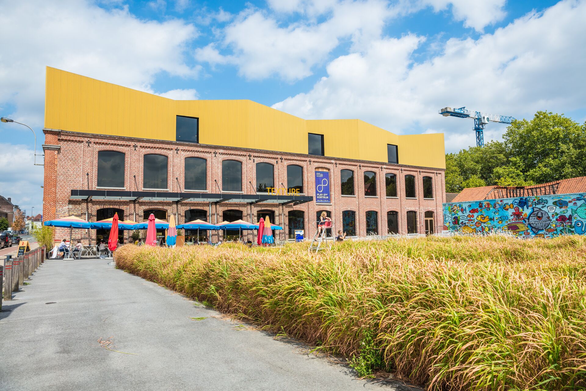 Texture, Kortrijk Industrieel gebouw van rood baksteen met een felgeel dak tegen een blauwe lucht, omgeven door hoog gras en kleurrijke muurschilderingen. In de buurt bieden parasols schaduw voor het buitenterras.