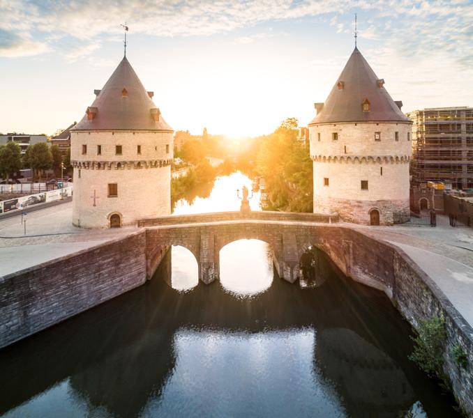 Twee middeleeuwse stenen torens flankeren een brug over een rustige rivier bij zonsopgang. De scène straalt rust uit, met zacht zonlicht dat op het water weerkaatst.