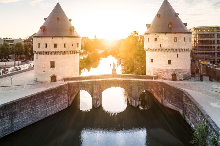Dos torres medievales de piedra flanquean un puente sobre un río tranquilo al amanecer. La escena irradia tranquilidad, con la suave luz del sol reflejándose en el agua.
