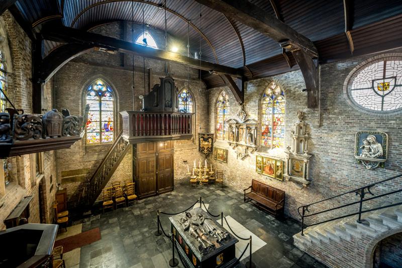 Ornate interior of a historic chapel with stained glass windows, a wooden staircase, and artwork on brick walls. Central tomb exudes solemnity and reverence.