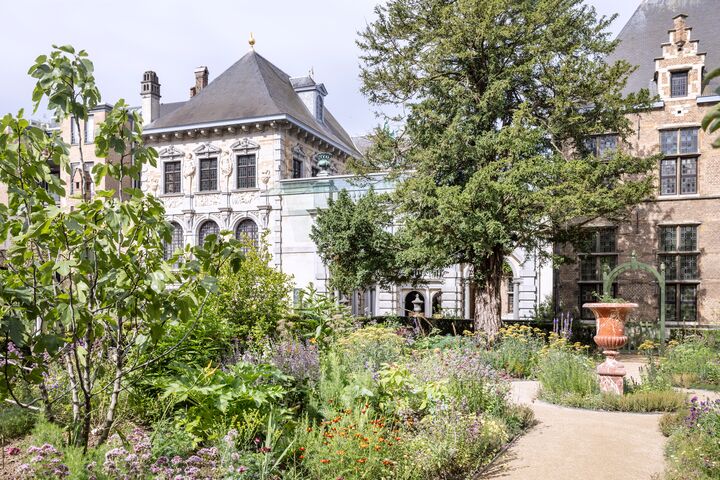 Een charmante tuin met weelderig groen en bloemen leidt naar een elegant, historisch gebouw. De scène voelt vredig en uitnodigend onder een heldere lucht.