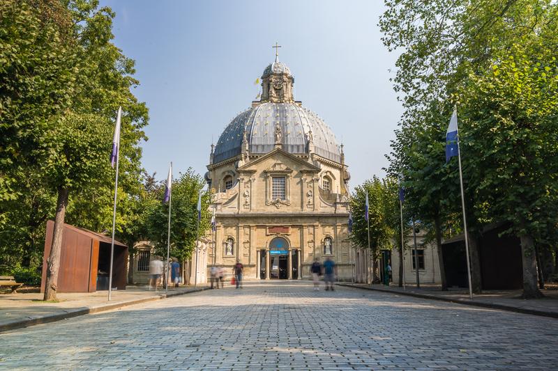 Historische koepelvormige kerk met gedetailleerde stenen gevel en kruis, omgeven door weelderige bomen, op een geplaveid plein. Mensen slenteren rustig rond en ervaren een serene sfeer.