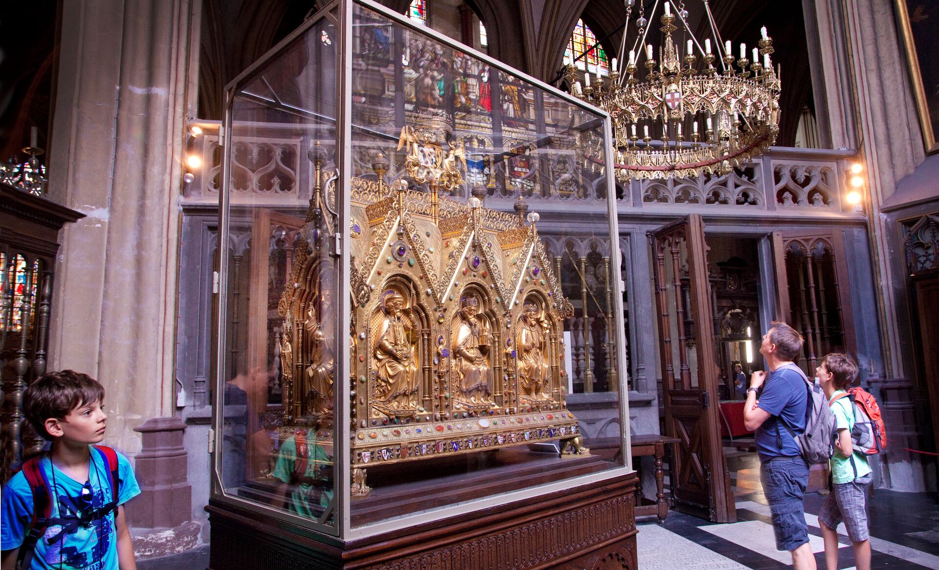 Bezoekers bewonderen een gouden, uitbundig versierd schrijn in het schemerige interieur van een kathedraal. De sfeer is plechtig, met ingewikkelde details en een grote kroonluchter boven.