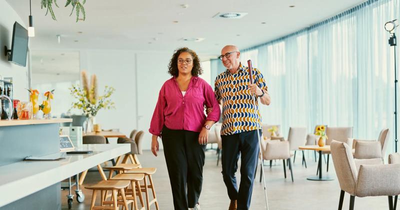 Twee mensen lopen door een lichte lounge met bar en stoelen; een man houdt een witte stok vast.