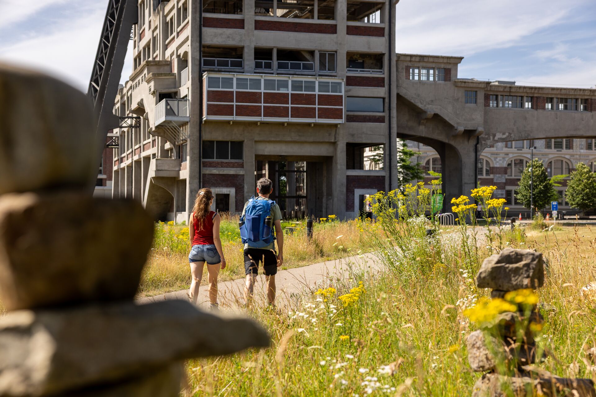 Escursionisti alla miniera di carbone di Waterschei Due escursionisti con zaini camminano nell'erba alta verso un grande edificio industriale in cemento.
