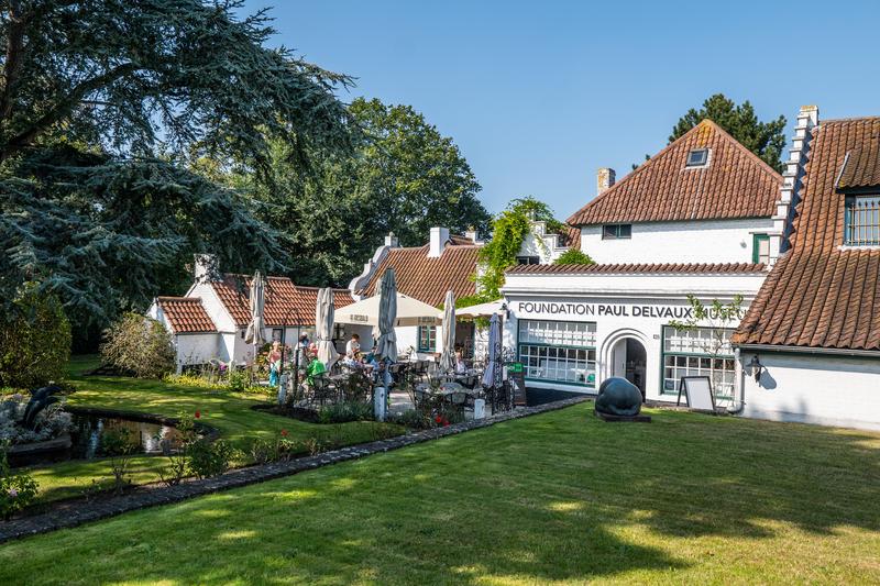 Een charmant wit gebouw met bruine dakpannen, met het opschrift "Foundation Paul Delvaux", omgeven door weelderig groen en een serene tuin onder een blauwe lucht.