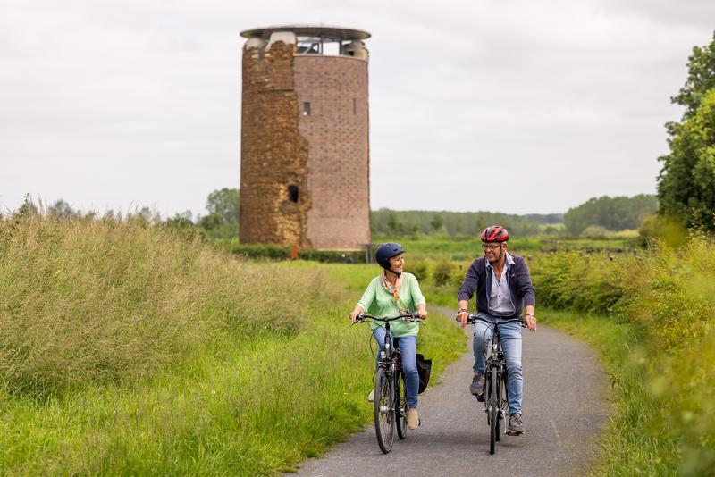 Een stel fietst over een landelijk pad, glimlachend en pratend. Een hoge, cilindervormige stenen toren en weelderig groen zijn zichtbaar onder een bewolkte lucht, wat een rustige sfeer oproept.