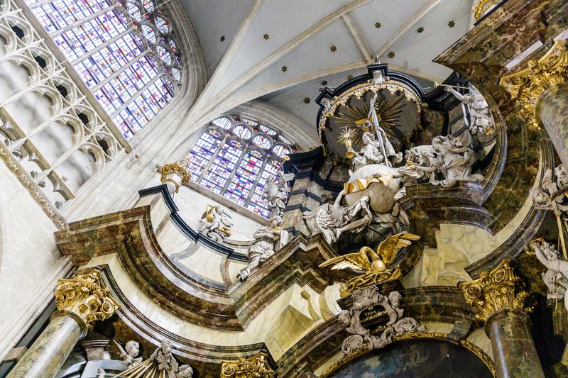 Intérieur d’église orné avec des sculptures détaillées et des accents dorés. Une grande fenêtre en vitrail projette une lumière colorée, créant une atmosphère majestueuse et respectueuse.