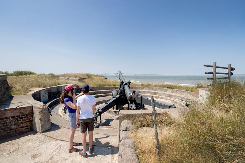Two children by a coastal bunker with a large cannon, looking out over sea and dunes under a blue sky.