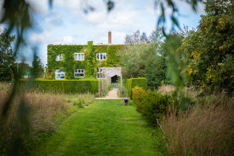 Een charmant tweeverdiepingshuis bedekt met weelderige klimop en een stenen boogvormige ingang. Het is omgeven door een goed onderhouden gazon, hoog gras en bomen onder een deels bewolkte lucht.