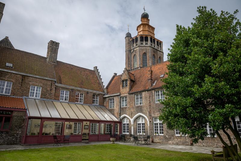 Historic brick building with red-tiled roofs, arch windows, and a prominent tower under a cloudy sky. Chairs and tables are in the grassy courtyard.