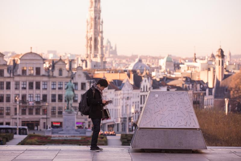 Man met rugzak leest op een heuvel met uitzicht op historische stad en hoge kerktoren in de verte.