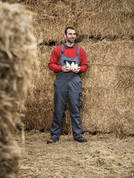 Thomas Cools Man in overalls and a red sweater stands in front of hay stacks and holds two chicks.