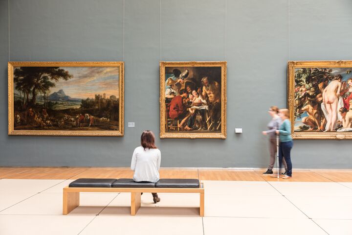 Museum room with visitors and paintings Museum room with three large paintings; one visitor sits on a bench, two others walk past.