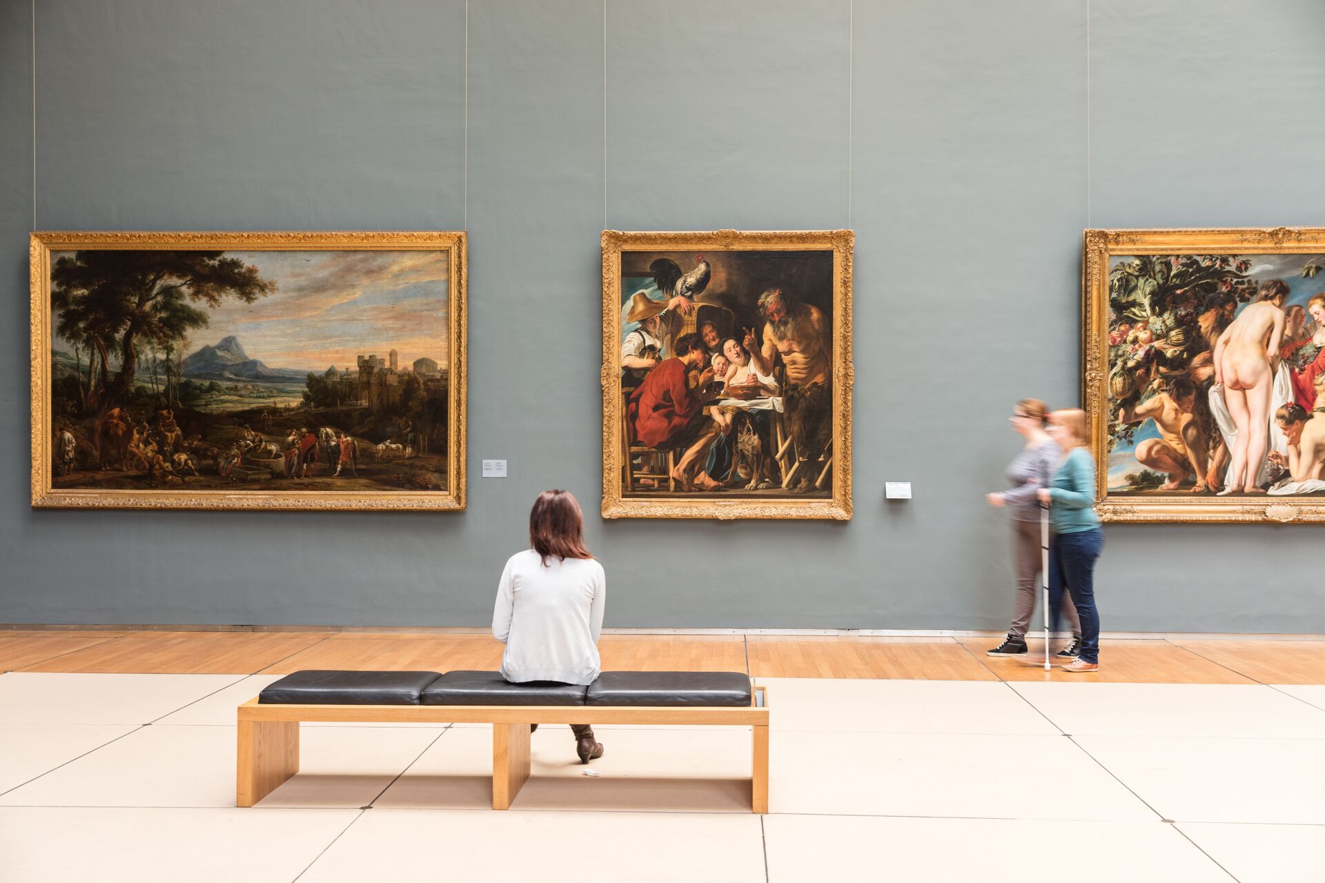 Museumzaal met bezoekers en schilderijen Museumzaal met drie grote schilderijen; een bezoeker zit op een bankje, twee anderen lopen langs.