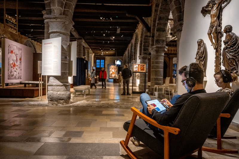 Museumzaal met bezoekers en audiogidsen, Museum Sint-Janshospitaal Museumzaal met stenen zuilen; bezoekers lopen rond terwijl twee mensen met koptelefoon in stoelen zitten.