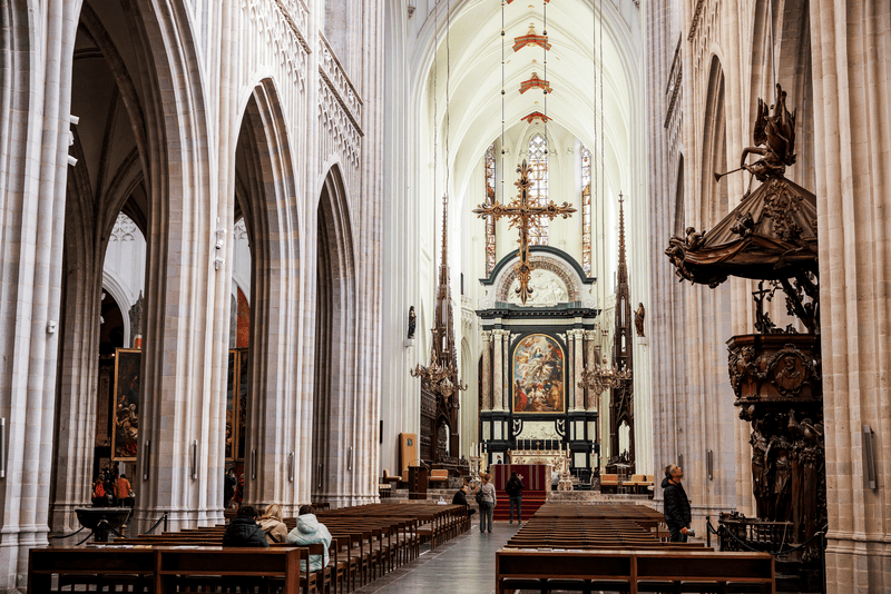Gotisch kathedraalinterieur met altaar in Antwerpen Interieur van een gotische kathedraal met hoge bogen, banken en altaar met kruis; enkele bezoekers lopen door het middenschip.