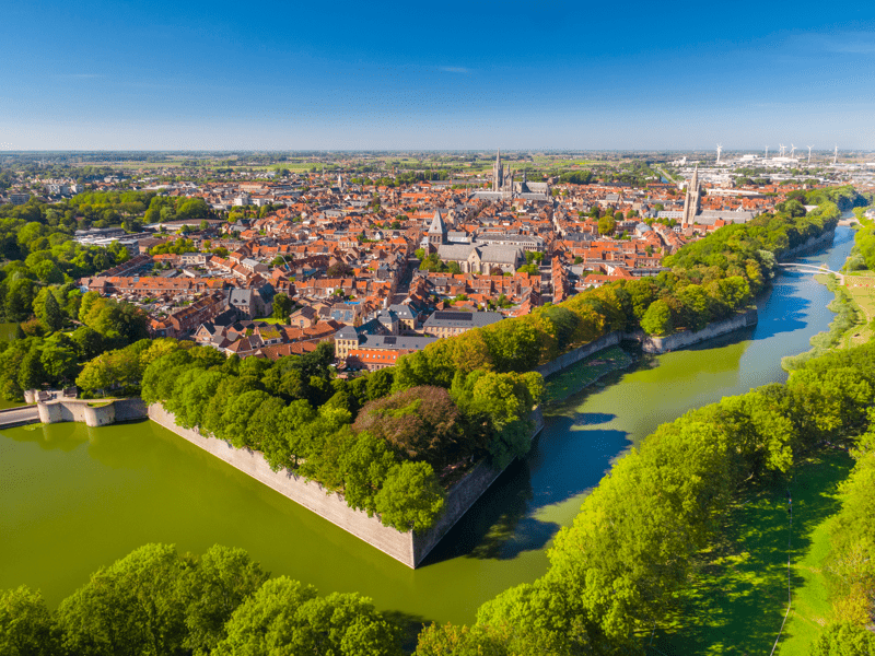 De Ieperse vestingen omringen de Belgische stad sinds de vroege middeleeuwen. Luchtzicht op een middeleeuwse stad met een rivier en dichte begroeiing eromheen. Roodachtige daken en kerktorens domineren het landschap onder een heldere blauwe lucht.