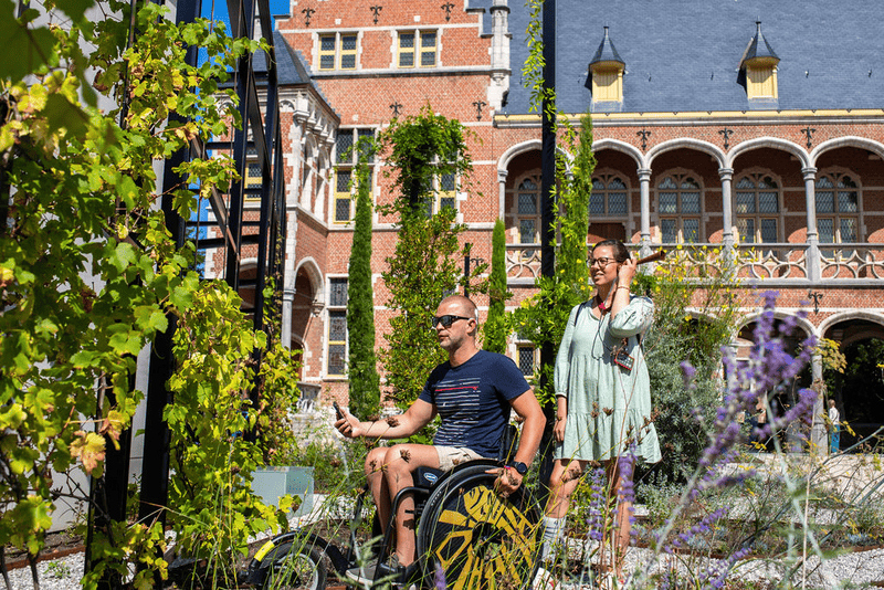 Bezoekers in tuin Hof van Busleyden Man in rolstoel en vrouw wandelen door groene tuin met klimop, met historisch rood bakstenen gebouw op de achtergrond.