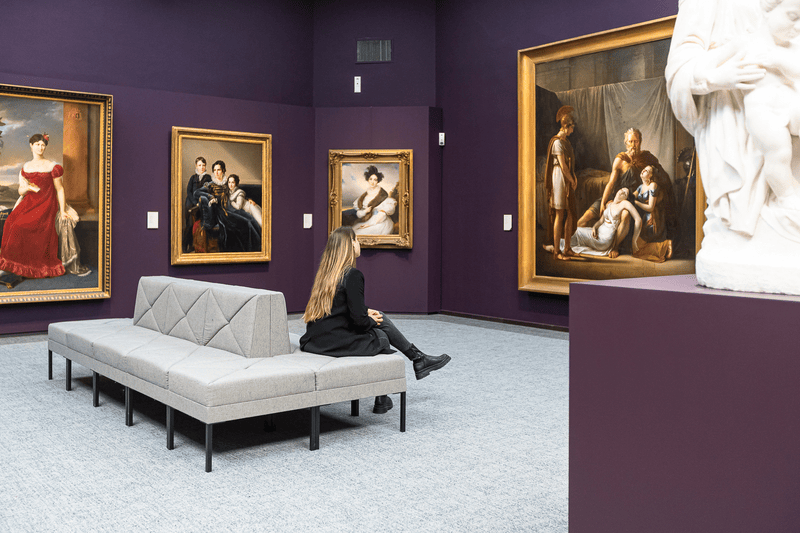 Museumzaal met schilderijen en zittende bezoeker, Groeningemuseum Bezoeker op bank in museumzaal met paarse muren, omringd door ingelijste schilderijen en een wit beeld.