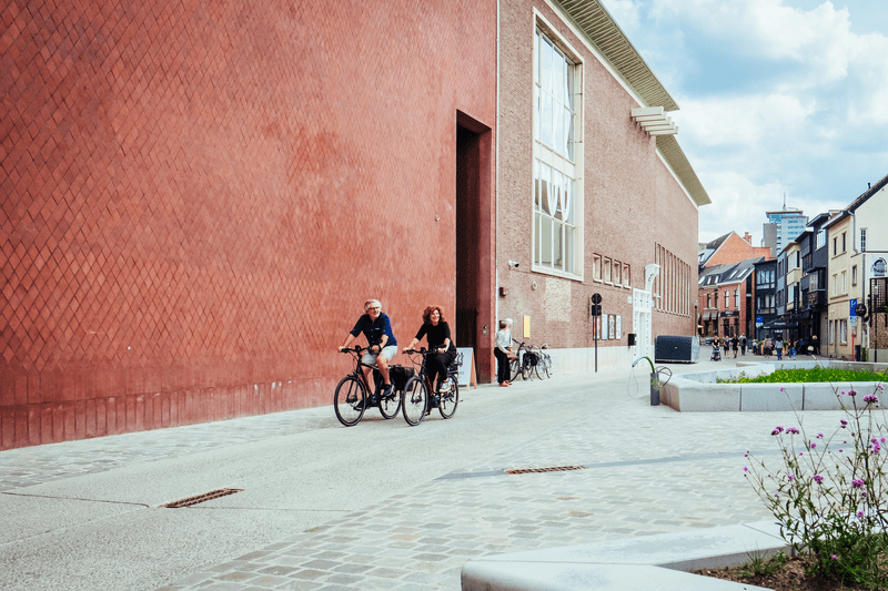 Two people are cycling side by side next to a large red brick building on a sunny day. The street is quiet, with modern and old buildings around