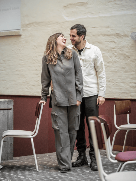 Ona Rombaut y Arne Breackman Pareja riéndose al aire libre, de pie entre sillas de café junto a una pared beige.