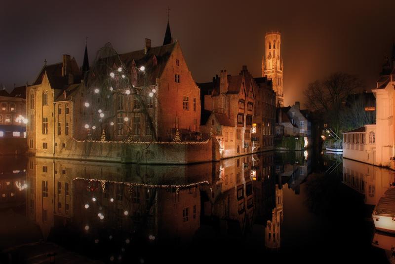 Historische bakstenen gebouwen weerspiegelen in een rustige gracht in Brugge ‘s nachts. Warme oranje lichten verlichten de scène en creëren een serene en magische sfeer.