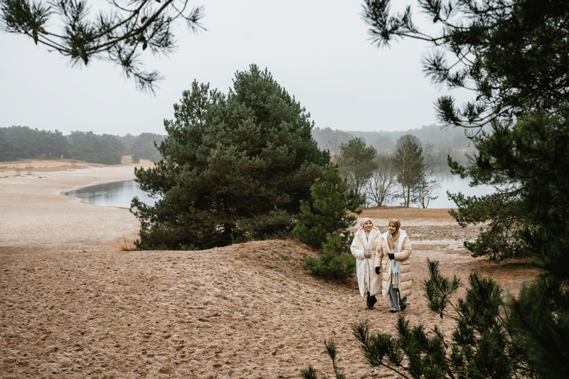 Bosland National Park Two people in warm coats walk through a sandy landscape with pine trees and a lake. The scene is tranquil and overcast, conveying a peaceful mood.
